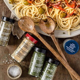 A styled flat-lay of Mediterranean-style pasta with vegetables on a plate, surrounded by jars of Ocho Río seasoning blends, two wooden spoons, coarse sea salt, and a blue lid on a rustic wooden surface.