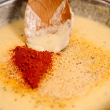 A close-up of creamy yellow cheese sauce being stirred with a wooden spoon, with a mound of red chili powder and other ground spices on top.