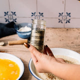 A hand with metallic gold nail polish holds a clear glass jar of Granulated Onion Seasoning over a rustic wooden table with baking ingredients.
