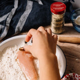 A hand with gold and black nail polish dips a raw chicken breast into a bowl of flour, with a jar of granulated onion seasoning and a wooden rolling pin in the background on a rustic wooden table.