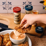 A hand with gold nail polish dips a piece of breaded chicken into a small bowl of creamy orange sauce. In the background, spice jars labeled 'Masala Chivda' and 'Masala Harisa' sit on a wooden cutting board next to a white plate with more breaded chicken.