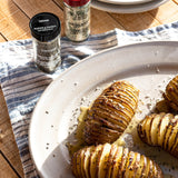 Sliced hassle potatoes on a white plate with herbs, next to spice jars labeled Thyme and Morton's Seasoning on a wooden table with a striped cloth.