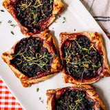 Four baked fruit tarts with flaky pastry crusts and dark berry果酱 centers, garnished with fresh thyme and orange zest, arranged on a white rectangular platter.