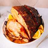 A glazed ham or roast served sliced in a white bowl with orange slices on a light tabletop.