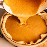 Pumpkin pie filling being poured from a white pot into a baked pie crust in a ceramic dish, shown in a close-up overhead view.