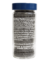 A clear plastic jar filled with dark brown or black sesame seed mixture, sealed with a dark blue lid, shown against a solid black background. The front label is turned sideways and displays the Nutrition Facts and product branding for Morton & Bassett Spice.