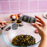 A person's hands are shown holding a glass jar of poppy seeds and pouring them into a gold measuring spoon over a large glass bowl filled with black and white poppy seeds, set against a pink tiled background.
