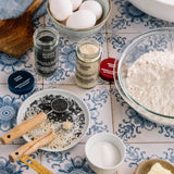 A styled flat-lay of baking ingredients on a blue-and-white patterned tile surface, including a bowl of flour, eggs, sea salt, poppy seed, and other spices, with measuring cups and utensils.