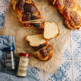 Braided poppy seed and sesame seed bread rolls topped with sesame seeds, arranged on parchment paper with slices cut out. Two spice jars labeled 'Poppy Seeds' and 'Sesame Seed' by Gordon & Basset are placed beside the bread on a blue patterned tile surface with a blue cloth.