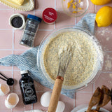 A top-down styled scene of a glass bowl filled with creamy poppy seed batter and a wooden-handled whisk, surrounded by baking ingredients on a pink tiled surface: vanilla extract, poppy seeds, lemon zest, a red lemon pie spice jar, eggshells, eggs, wooden spoons, and a blue cloth.