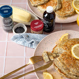 A styled breakfast scene on a pink tiled table with plates of pancakes topped with lemon slices, a bottle of vanilla syrup, a jar of lemon poppy seed spice, and a bottle of poppy seed flour.