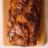 A freshly baked loaf of bread with a glossy, dark brown crust sits on a light-colored wooden cutting board.