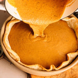 Pumpkin pie filling being poured from a white pot into a baked pie crust in a ceramic pie dish.