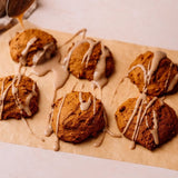 Six golden-brown pumpkin spice cookies drizzled with light beige icing, arranged on parchment paper with a spoon pouring more icing in the background.