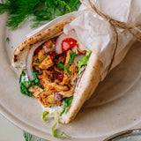 A close-up of a stuffed pita wrap tied with twine, filled with grilled chicken, red onion, tomato, dill, and white sauce, served on a beige plate.