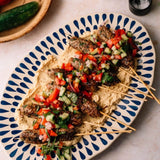 Greek kebab skewers topped with diced red pepper, cucumber, and onions over a bed of hummus, served on a blue and white patterned oval platter.