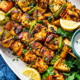 Grilled chicken tikka skewers on a white serving platter, garnished with chopped cilantro, served with lemon wedges and a small bowl of white chutney.