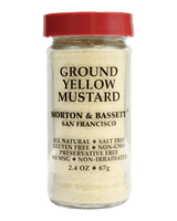 A jar of Morton & Bassett Ground Yellow Mustard against a solid black background. The clear glass jar shows the pale yellow mustard powder inside, topped with a red lid and featuring black text on the front label.