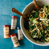 Four spice jars arranged on a blue surface beside a white bowl of mixed greens and shredded cabbage with wooden spoons. Visible labels include Garlic Powder, Ground Turmeric Mushroom, and Ground Black Pepper.