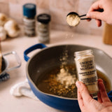 A hand holds a jar of Morton & Basset Ground Yellow mustard in front of a blue pot on a kitchen counter, with another hand sprinkling mustard powder over the oil. Garlic and other spices are blurred in the background.