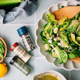 A scalloped white bowl filled with a green salad containing cucumbers, crumbled cheese, and a wooden spoon, styled on a light blue tile surface with three spice jars, a lemon, and other ingredients nearby.