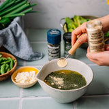 A person's hands are shown adding ground yellow mustard from a glass jar into a bowl of green herbs and oil, with various fresh ingredients like green beans and onions in the background on a tiled countertop.