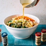 A large white bowl filled with mixed vegetables and shredded carrots, with a hand pouring a yellow dressing from a small white pitcher. Seasoning bottles sit on a blue tiled surface against a light speckled wall.