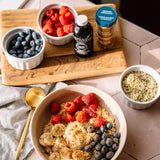 A styled breakfast scene with a large bowl of oatmeal topped with strawberries, blueberries, banana slices, and granola, alongside small bowls of blueberries, raspberries, vanilla extract, and ground cinnamon on a wooden board.