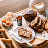 Slices of chocolate chip bread on parchment paper with a plate, knife, vanilla extract bottle, and eggs on a wooden table.