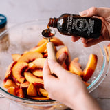 A person's hands pour dark brown WBH Pure Vanilla Extract from a small bottle into a measuring spoon over sliced peaches in a glass bowl.