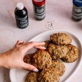 A hand reaches for a golden-brown baked cookie on a white oval plate, with three bottles of MFP vanilla extract flavors in the background on a light pink marble surface.