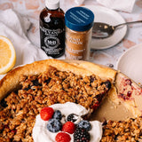A baked berry pie or crumble dessert in a round dish, topped with whipped cream and fresh berries, with vanilla extract and cinnamon powder bottles behind it on a light kitchen-style surface.