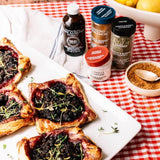 Four baked tartlets topped with dark berries and thyme are arranged on a white rectangular platter, with M&B Gourmet food ingredients including almond extract, ground lemon, thyme, vanilla powder, and cinnamon scattered on a red-and-white checkered blanket.