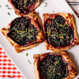 Four baked fruit tarts with golden flaky pastry crusts, dark berry果餡, and fresh thyme garnish, arranged on a white rectangular serving platter over a red-and-white checkered cloth.