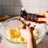 A person's hands with red nail polish are shown pouring a clear liquid from a dark amber bottle labeled 'Purified Stevia' into a gold measuring spoon over a glass bowl containing pale yellow dough or batter, set against a tiled kitchen background.