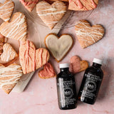 Two bottles of M&B Beauty Pure Vanilla & Almond Extract makeup removers arranged on a pink marble surface with heart-shaped cookies and cookies with drizzles of white and pink icing around them.