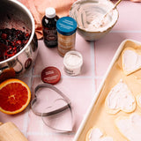 A styled flat-lay of baking ingredients and tools on a pink tiled surface, including a heart-shaped metal cutter, vanilla extract, ground cinnamon, powdered sugar, a bowl of white frosting, and heart-shaped cookies on a baking sheet.