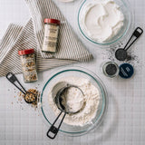 Overhead flat-lay of baking ingredients on a white grid surface: a large glass bowl with flour and measuring cups, a smaller bowl with white cream, two spice jars labeled Sesame Seed and Flaxseed, measuring spoons, and scattered seeds and spices.