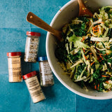 Four spice jars arranged on a blue surface beside a large white bowl of mixed greens and shredded cabbage with wooden spoons. The labels visible include Garlic Powder, Ground Yellow Mustard, Ground Black Pepper, and Onion & Garlic Seasoning.
