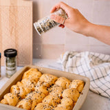 A hand sprinkles black sesame seeds and white sea salt over freshly baked cinnamon rolls in a beige baking pan. The scene is set on a kitchen counter with a checkered kitchen towel and another spice jar in the background.