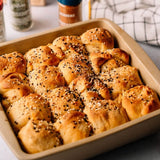 Baked bread rolls or buns topped with sesame and poppy seeds, arranged in a beige rectangular baking dish on a light surface with blurred spices and a checkered cloth in the background.