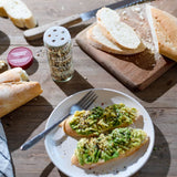 Avocado toast on a white plate with a fork, set on a rustic wooden table next to a jar of seasoning, sliced bread on a cutting board, and more bread pieces.