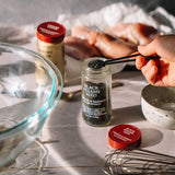 A hand holding tweezers over a glass jar of Black Sesame Seed seasoning, with raw chicken pieces and another jar of seasoning in the background on a kitchen counter.