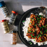 A top-down styled food scene showing a black skillet with chicken and rice on a tiled surface, alongside several spice bottles including Wicked Pepper, Black Sesame Seed, Ground Ginger, and Beef Calla Lilies.