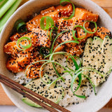 A close-up overhead shot of a bowl of salmon sushi rice bowl, featuring orange salmon pieces over white rice, topped with sesame seeds, sliced avocado, and green onion, with chopsticks resting at the bottom left.