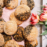 A close-up overhead view of several round sesame-topped bread rolls arranged on a light pink cloth beside pink tulips. The rolls are golden brown with white sesame seeds on some and black sesame seeds on others.