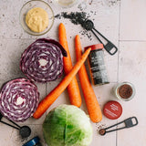 Overhead flat-lay of cooking ingredients on a light stone surface: two halves of red cabbage, a whole green cabbage, four carrots, a bottle of black sesame seeds, measuring spoons, and small bowls of mustard and seasoning.