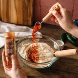 A person's hands are shown seasoning a bowl of dry ingredients with smoked paprika from a glass jar. The scene is set on a rustic wooden table with a cutting board and kitchen tiles in the background.