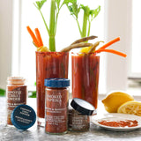 Two Bloody Mary cocktail glasses filled with red drink and garnished with celery, carrots, and lemon, styled with jars of Morton & Bassett spices including Chipotle Chili Powder, Smoked Paprika, and Lemon Pepper Blend, along with lemons and a plate of red seasoning on a countertop near a window.