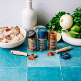 Three jars of Morton and Bassett spices arranged on a blue tiled surface with raw shrimp, onion, cilantro, lime, and a white bottle in the background.